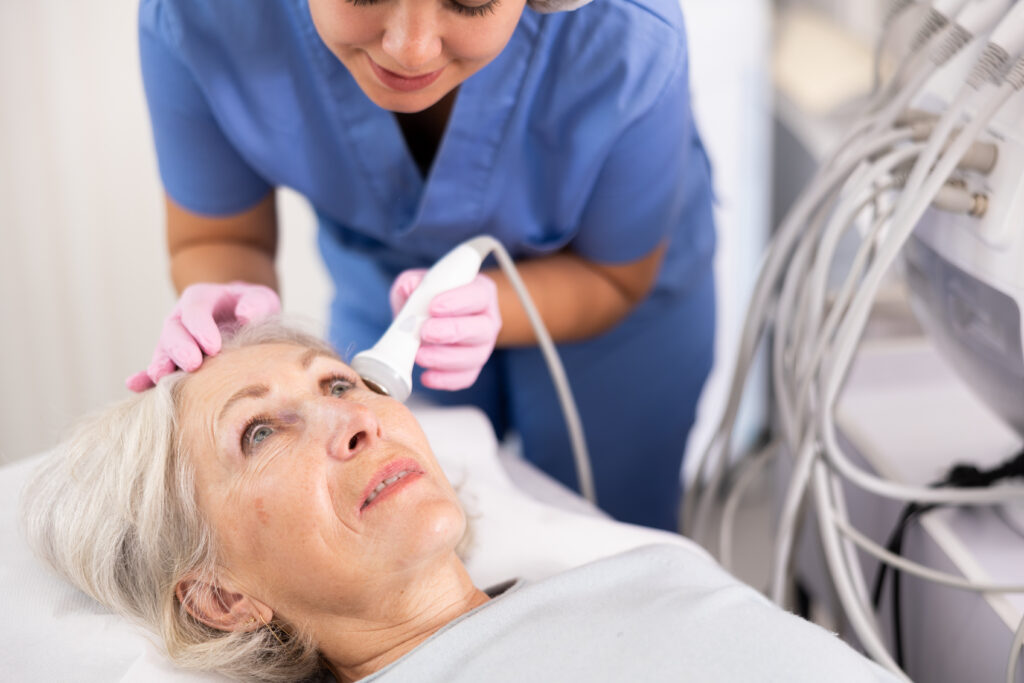 Delighted old woman lying on clinical chair in aesthetic cabinet during face laser therapy procedure